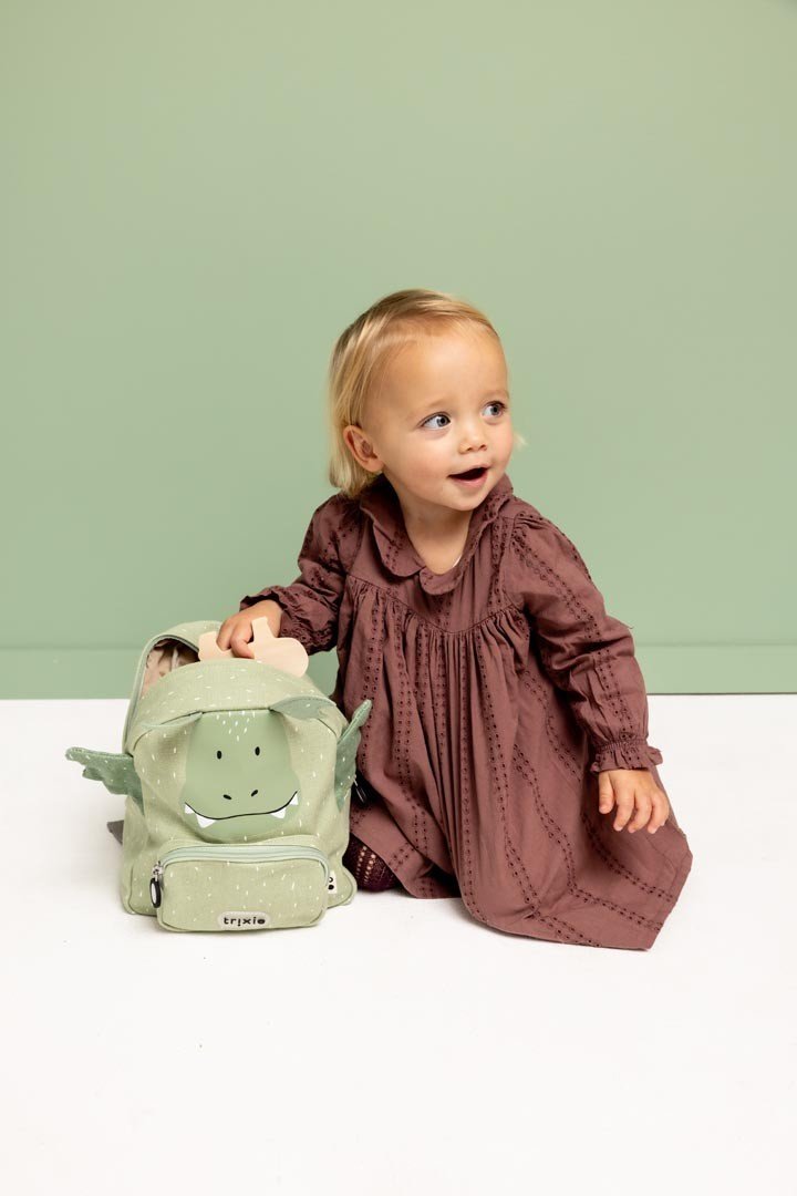 9. Child sitting next to green dragon-themed backpack, wearing a brown dress