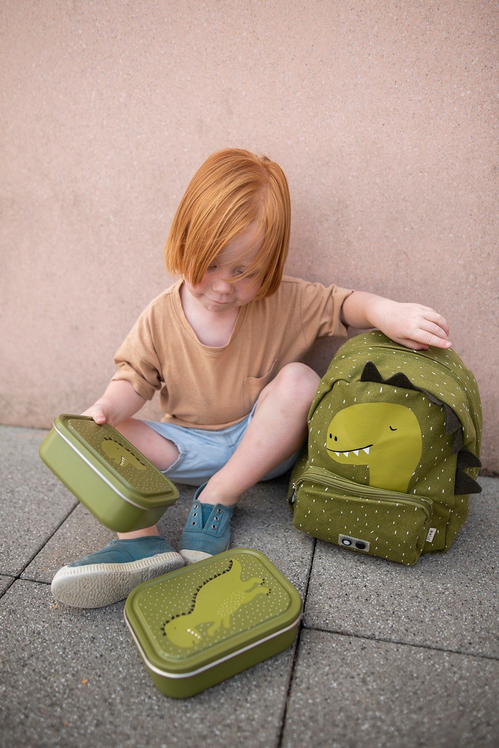 1. Child sitting on pavement with green Mr. Dinosaur lunch box and matching backpack, showcasing playful design