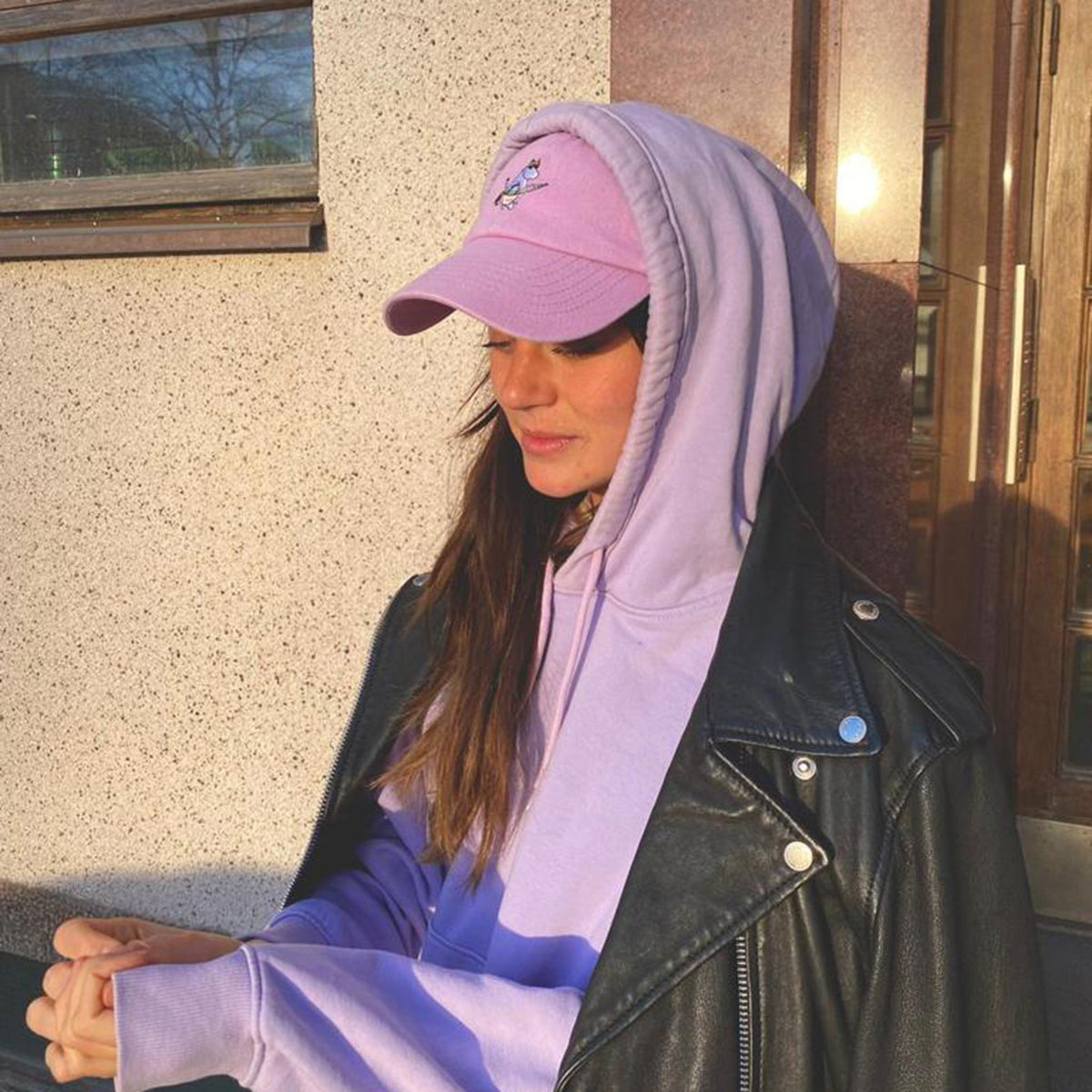 1. Woman wearing pink Snorkmaiden Dad Cap by Moomin with purple hoodie and black leather jacket outdoors