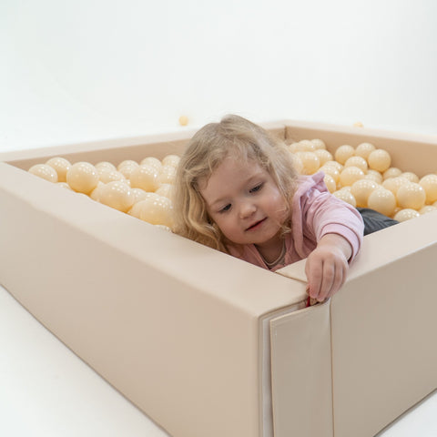 1. Young girl playing in a beige ball pit filled with soft play balls, wearing a pink top, in a playful setting