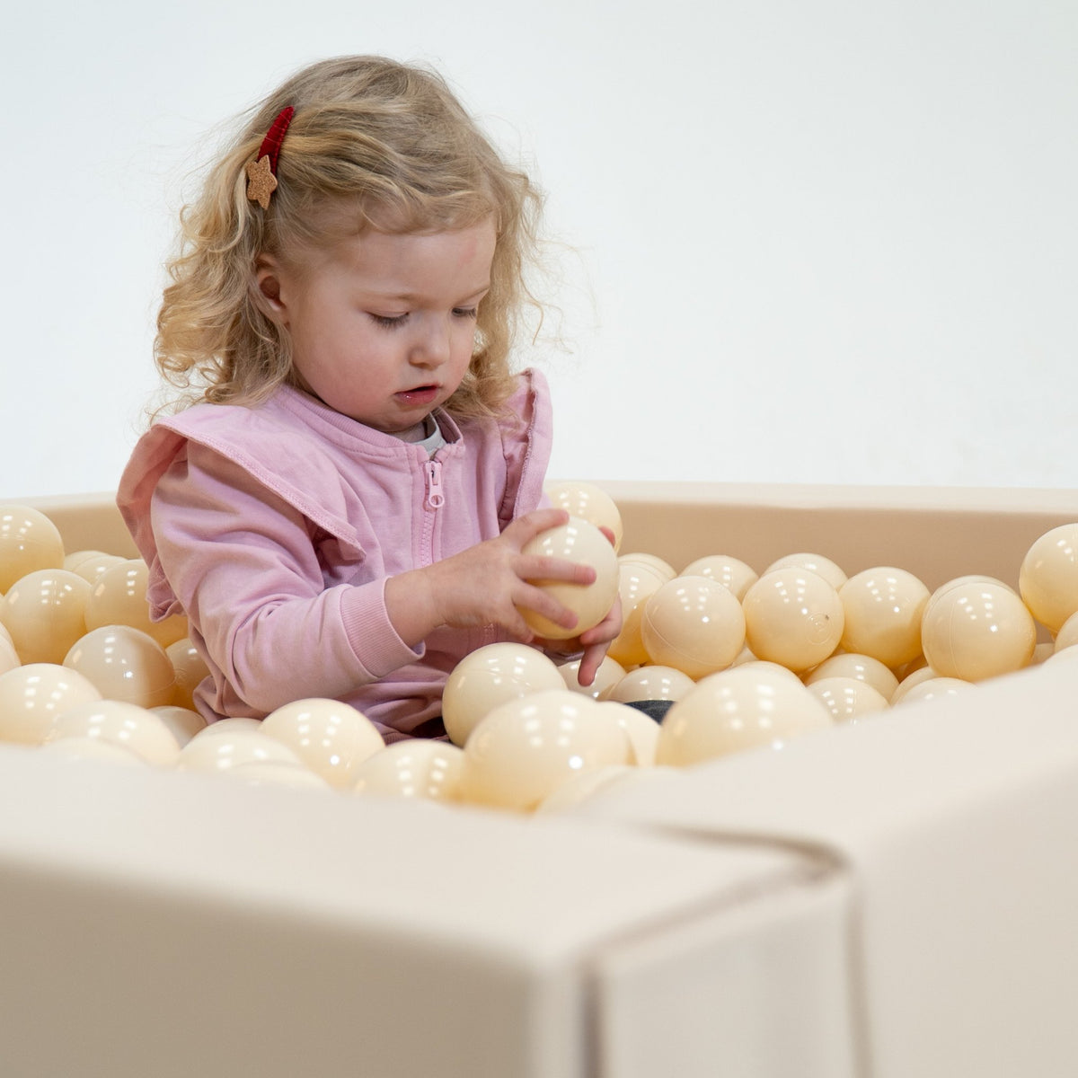 1. Young girl in a pink outfit sitting in a beige ball pit, holding a soft play ball, engaging in sensory play