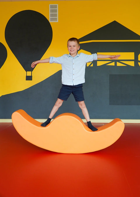 1. Boy standing on orange IGLU Soft Play Double Rocker Buddy in playroom with colorful wall mural