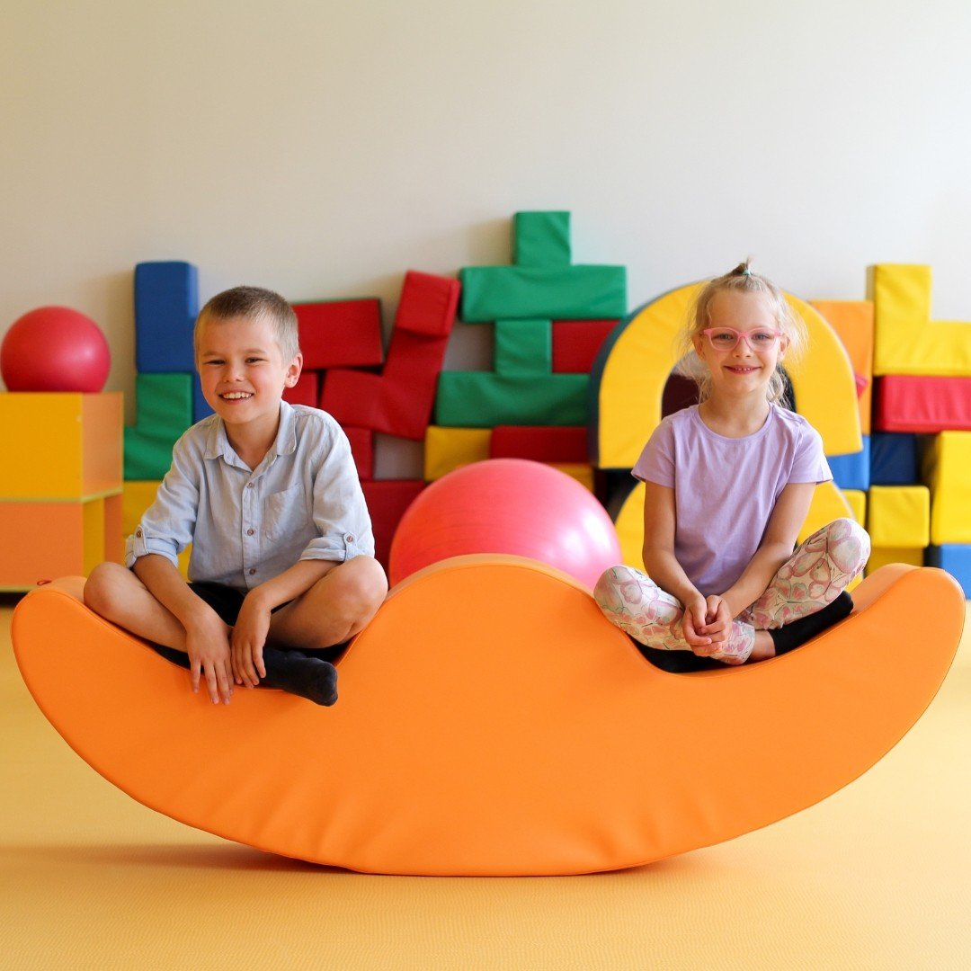 1. Two children sitting on orange soft play double rocker in playroom with colorful blocks, smiling and enjoying playtime