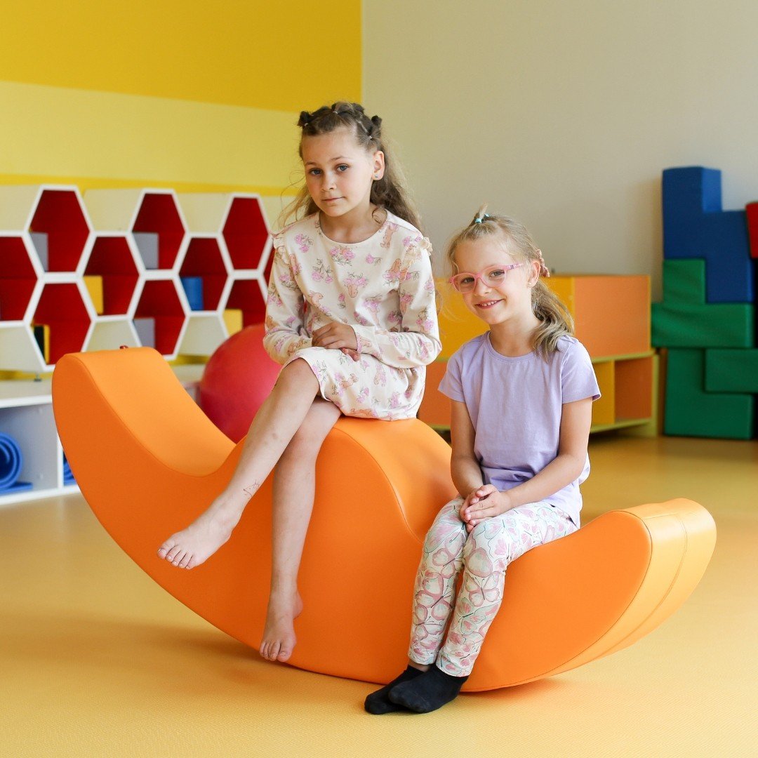 6. Two girls sitting on orange IGLU Soft Play Double Rocker Buddy in playroom with hexagonal shelves