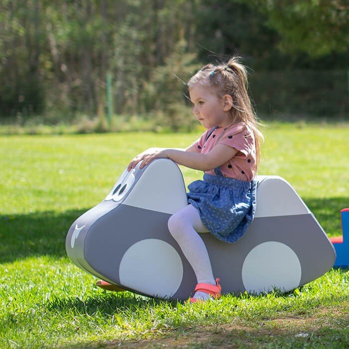 3. Girl in pink top and blue skirt riding IGLU grey car-shaped soft play toy on grass, enjoying outdoor play