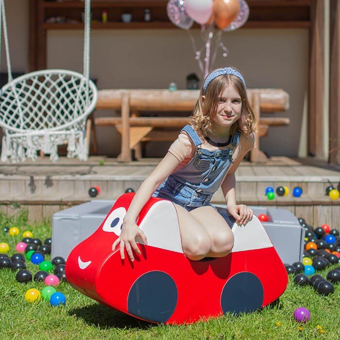 10. Girl sitting on IGLU red soft play ride-on car toy in a garden setting with colorful balls