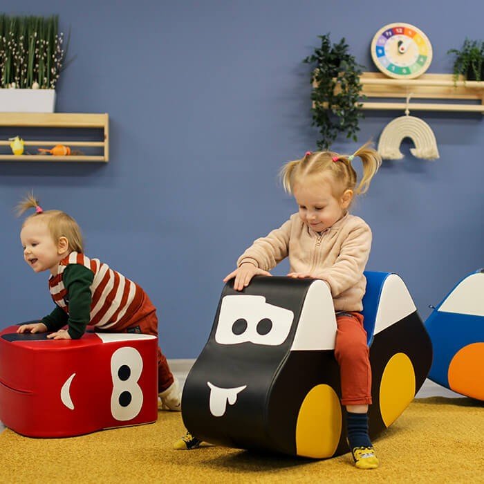 6. Two toddlers playing with IGLU soft play ride-on toys, one red and one black car, in a playroom
