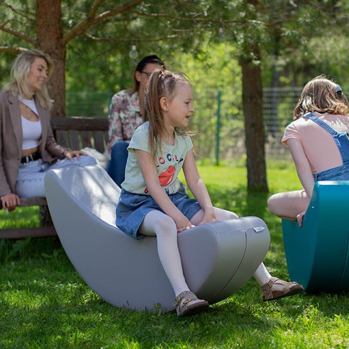 1. Young girl sitting on grey banana-shaped rocking toy outdoors with adults in background