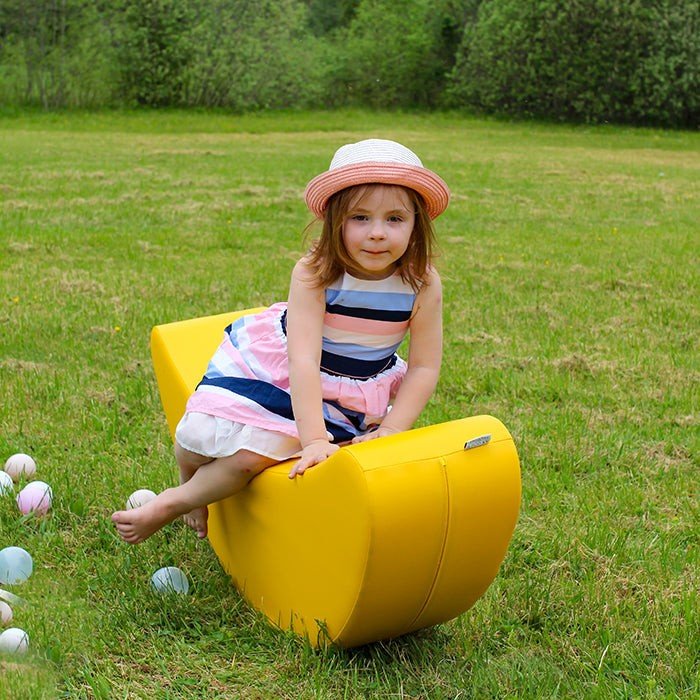 1. Young girl in striped dress and hat sitting on yellow banana-shaped rocking toy in grassy field