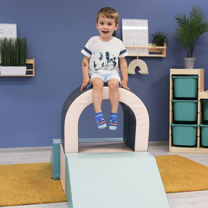 1. Boy sitting on top of IGLU pastel soft play tunnel set in indoor playroom with blue wall