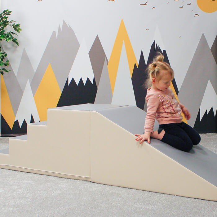 7. Child sliding down beige and grey soft play set in a playroom with mountain wall art