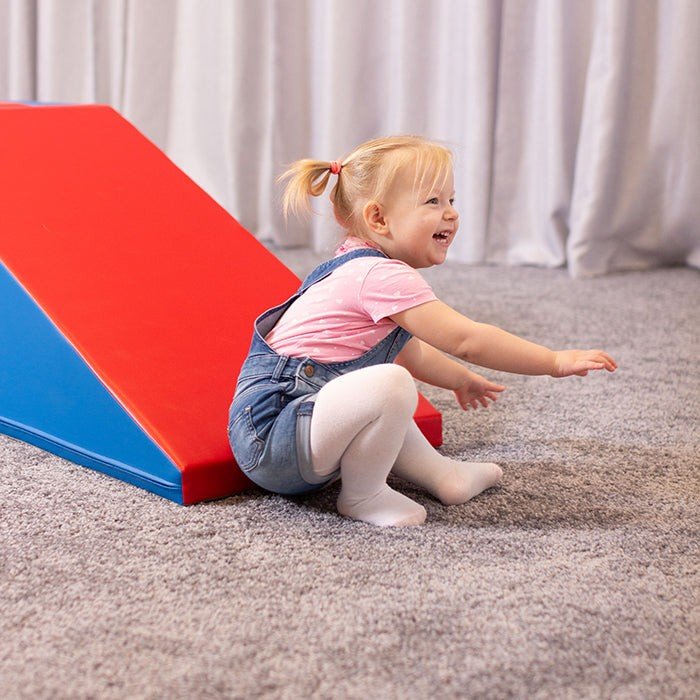 1. Smiling toddler sitting on blue and red soft play slide in a carpeted room