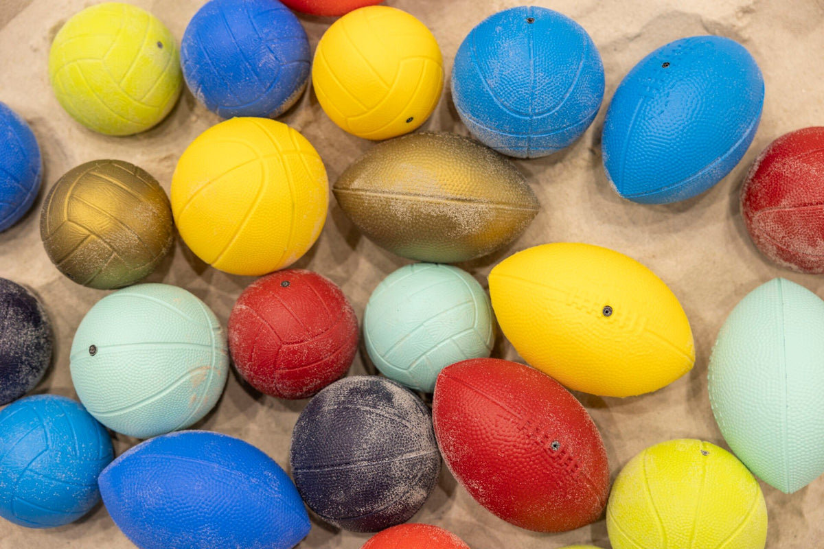 4. Close-up of various colorful sports balls on sand, including rugby, basketball, volleyball, and football