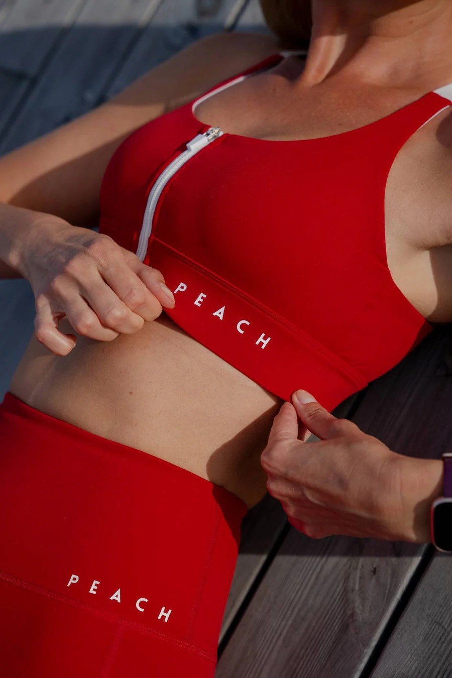 1. Woman adjusting Peachlabel 'Cute Cranberry' red sports bra with front-zip, showing supportive elastic band