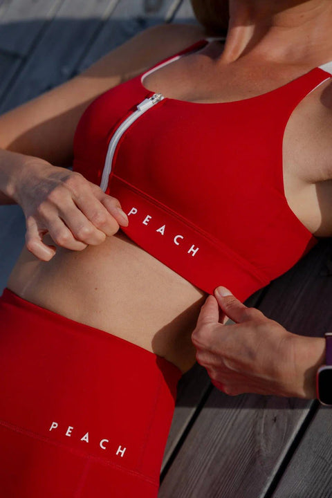 1. Woman adjusting Peachlabel 'Cute Cranberry' red sports bra with front-zip, showing supportive elastic band