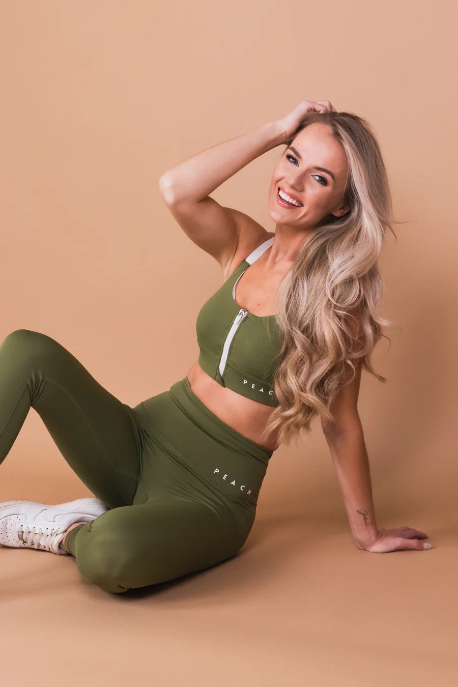 2. Woman sitting in Peachlabel Olive Green Sports Bra and leggings, showcasing front-zip design, studio setting