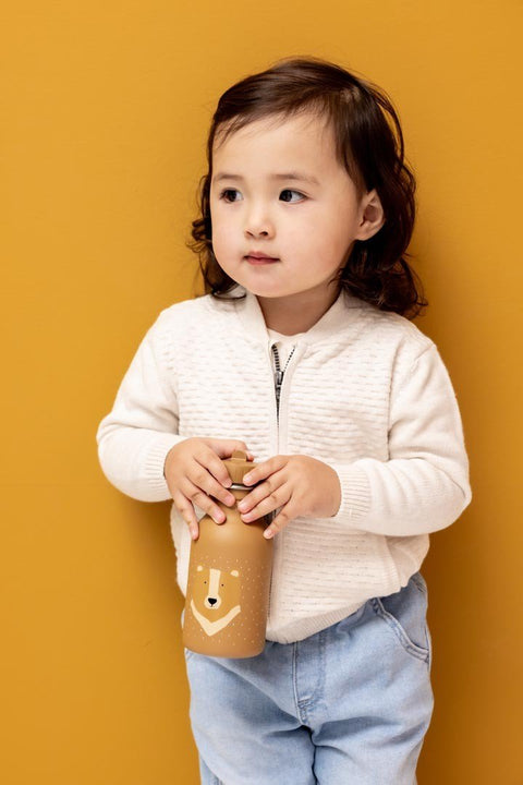 1. Child holding Trixie Baby Mr. Bear stainless steel bottle, wearing beige sweater and jeans, against mustard background