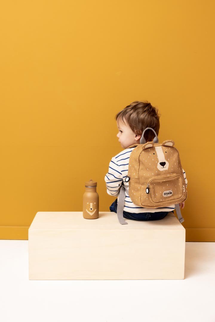 1. Child sitting with Trixie Baby Mr. Bear backpack and stainless steel bottle, wearing striped shirt, against mustard background