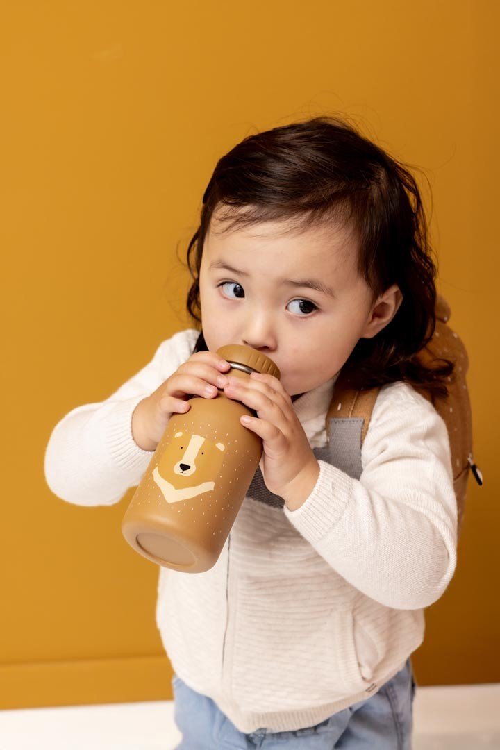 1. Child holding Trixie Baby Mr. Bear stainless steel bottle, wearing beige sweater, against mustard background
