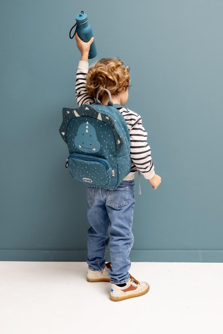 4. Child with backpack holding blue Mr. Triceratops bottle, wearing striped shirt and jeans, against blue wall