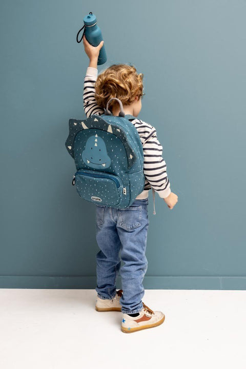4. Child with backpack holding blue Mr. Triceratops bottle, wearing striped shirt and jeans, against blue wall