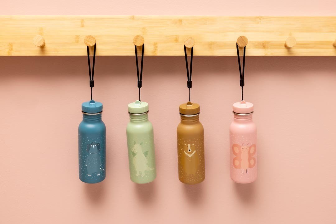 7. Four Trixie Baby stainless steel bottles in blue, green, brown, and pink hanging on hooks, against pink background