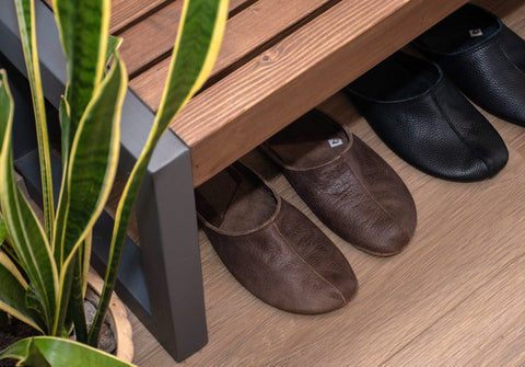 1. Dark brown unisex leather slippers under wooden bench beside potted plant on wooden floor