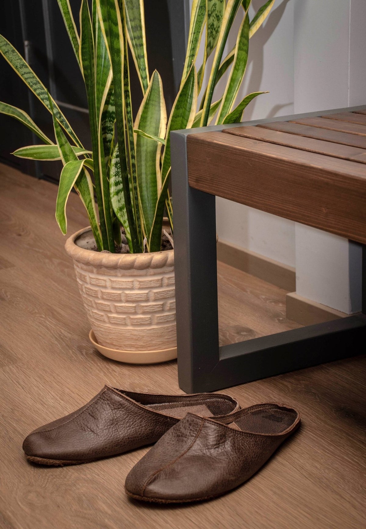 1. Pair of dark brown unisex leather slippers placed on wooden floor next to potted plant and bench