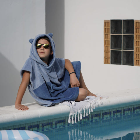 1. Child wearing blue bath poncho with hood and ears, sitting by a pool, wearing sunglasses, in a sunny outdoor setting