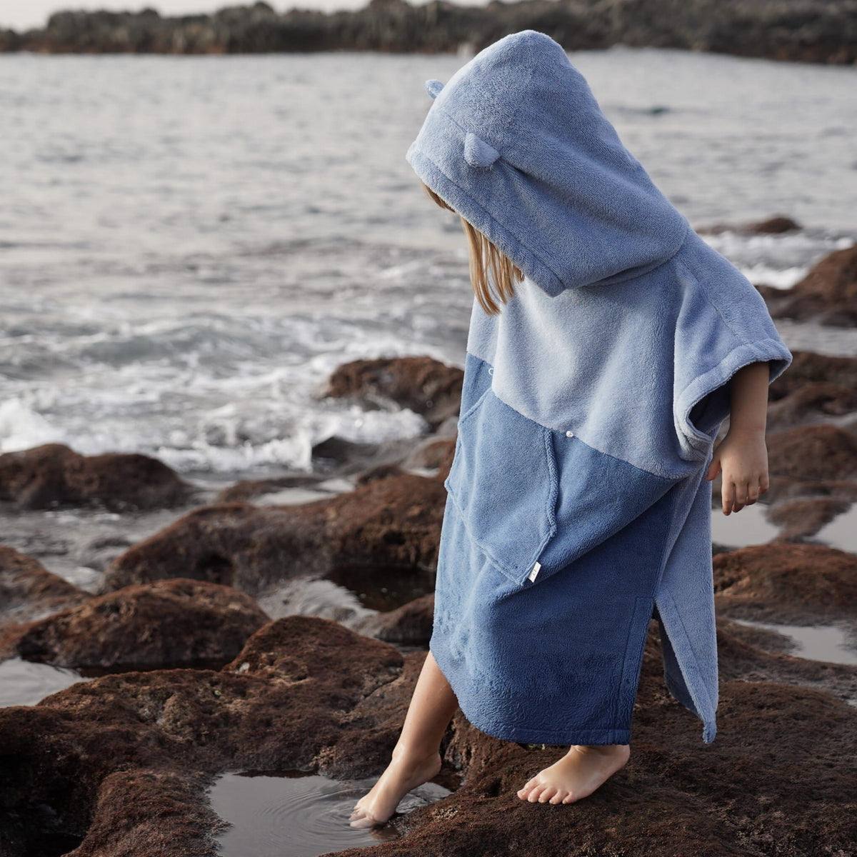 1. Child wearing blue bath poncho with hood and ears, walking on rocky beach, looking down at water