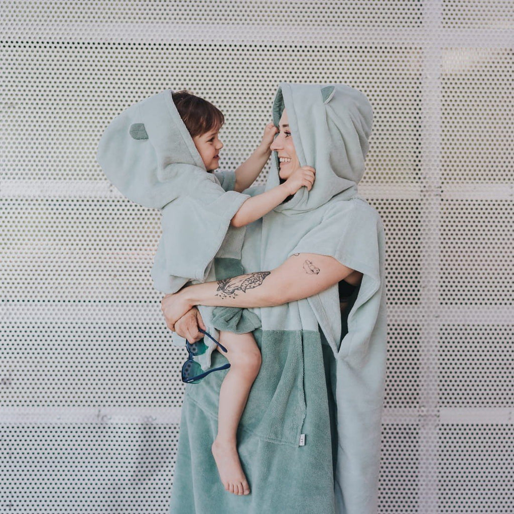 1. Adult and child wearing matching green Surfbear bath ponchos with hoods and ears, smiling at each other, in front of a metal background