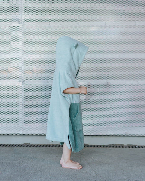 1. Side view of child wearing green Surfbear kids bath poncho with hood and ears, standing in front of a metal background