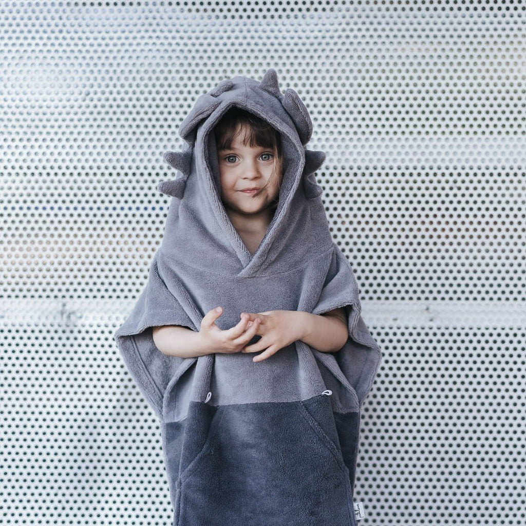 1. Child wearing grey Surfdragon kids bath poncho with hood and ears, standing against a perforated metal background