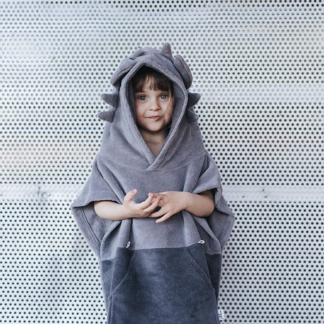 1. Child wearing grey Surfdragon kids bath poncho with hood and ears, standing against a perforated metal background