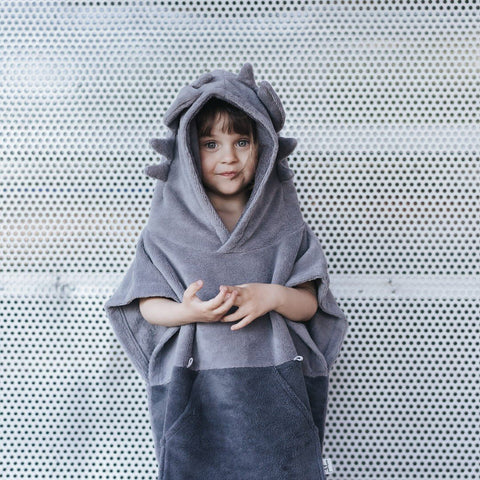 1. Child wearing grey Surfdragon kids bath poncho with hood and ears, standing against a perforated metal background