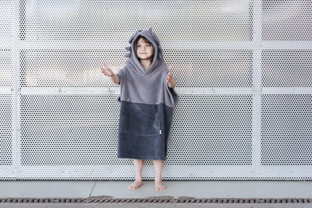 4. Child wearing grey Surfdragon kids bath poncho with hood and ears, standing with arms outstretched against a perforated metal background