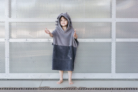 4. Child wearing grey Surfdragon kids bath poncho with hood and ears, standing with arms outstretched against a perforated metal background