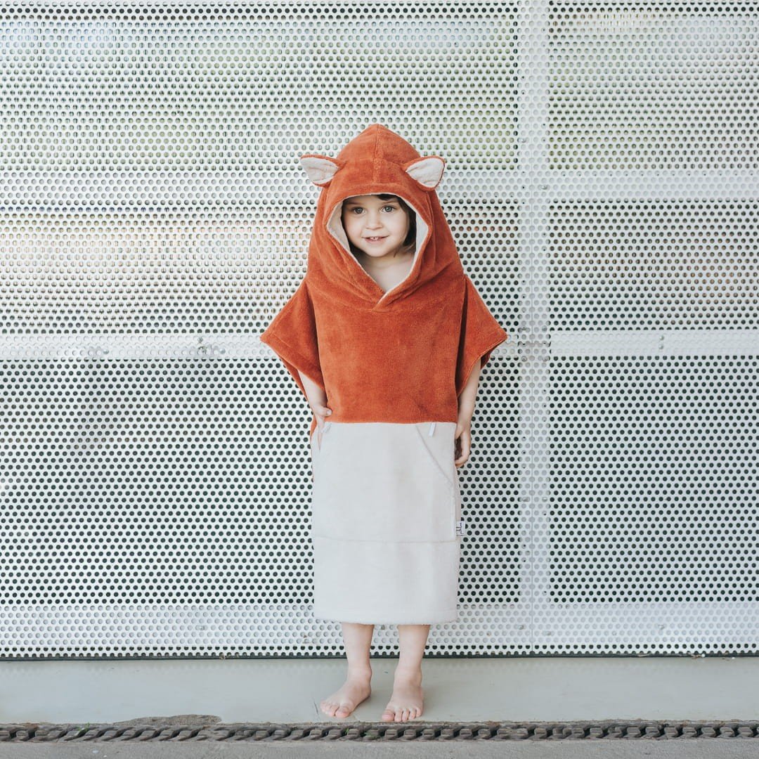 1. Child wearing orange Patulove Surffoxy kids bath poncho with hood and ears, standing against perforated metal background