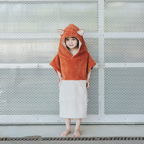 1. Child wearing orange Patulove Surffoxy kids bath poncho with hood and ears, standing against perforated metal background