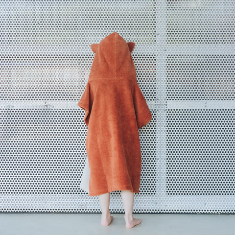 2. Back view of child wearing orange Patulove Surffoxy kids bath poncho with hood and ears against perforated metal background