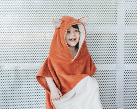 1. Child wearing orange Patulove Surffoxy kids bath poncho with hood and ears, smiling against a perforated metal background