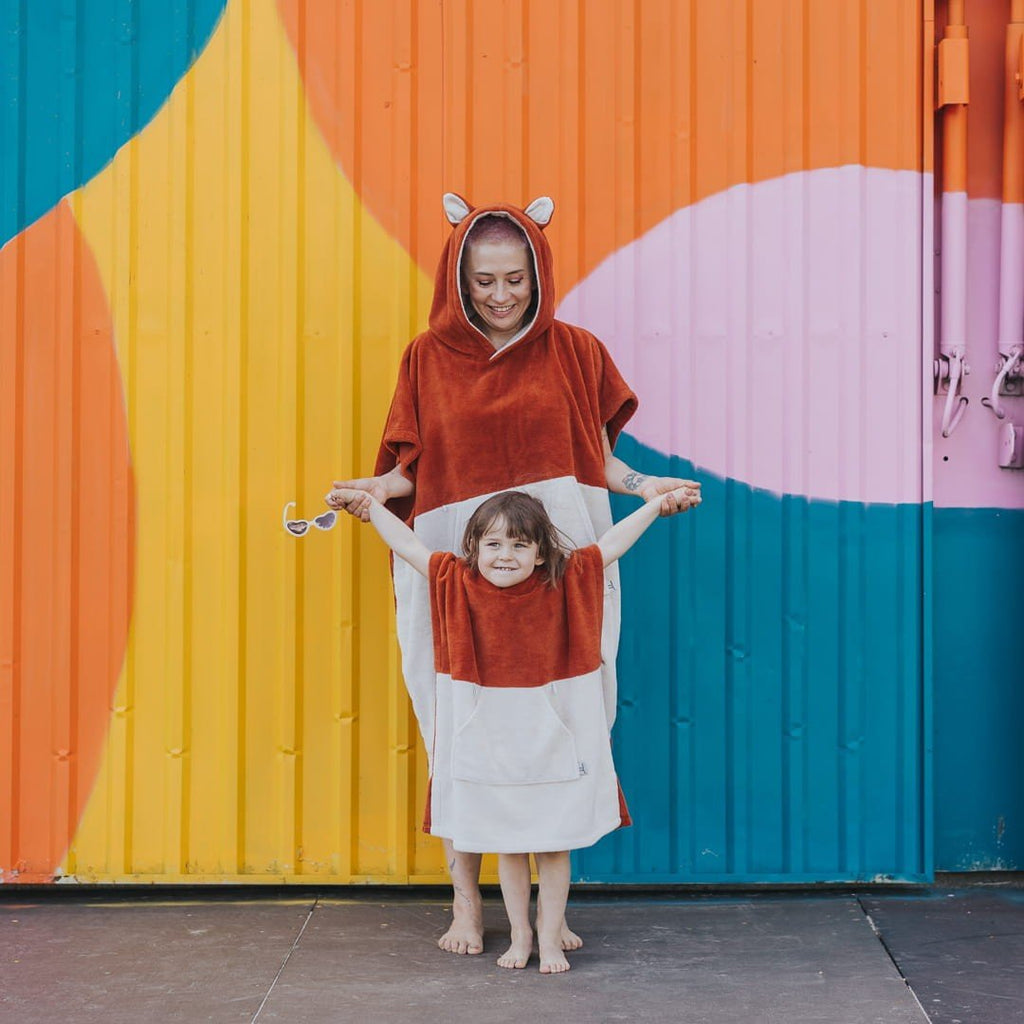 1. Adult and child wearing matching orange Patulove Surffoxy bath ponchos with hoods and ears, standing in front of colorful wall