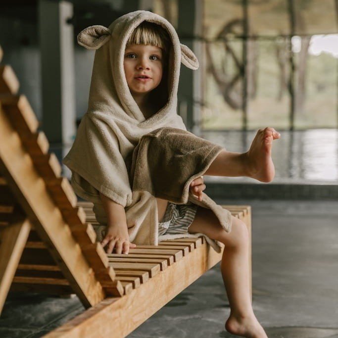 1. Child wearing beige Surfsheep kids bath poncho with hood and ears sitting on wooden lounge chair by indoor pool