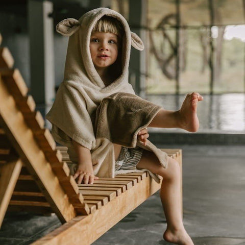 1. Child wearing beige Surfsheep kids bath poncho with hood and ears sitting on wooden lounge chair by indoor pool