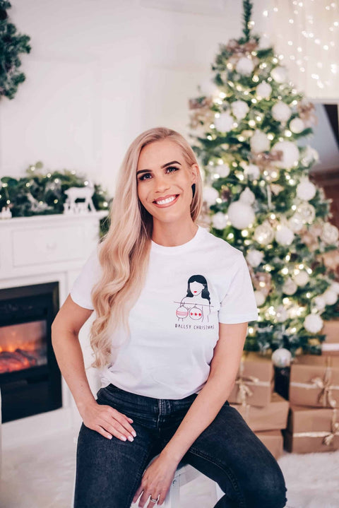 1. Woman sitting in festive room wearing white 'Ballsy Christmas' T-shirt with illustration, paired with jeans, in front of decorated Christmas tree