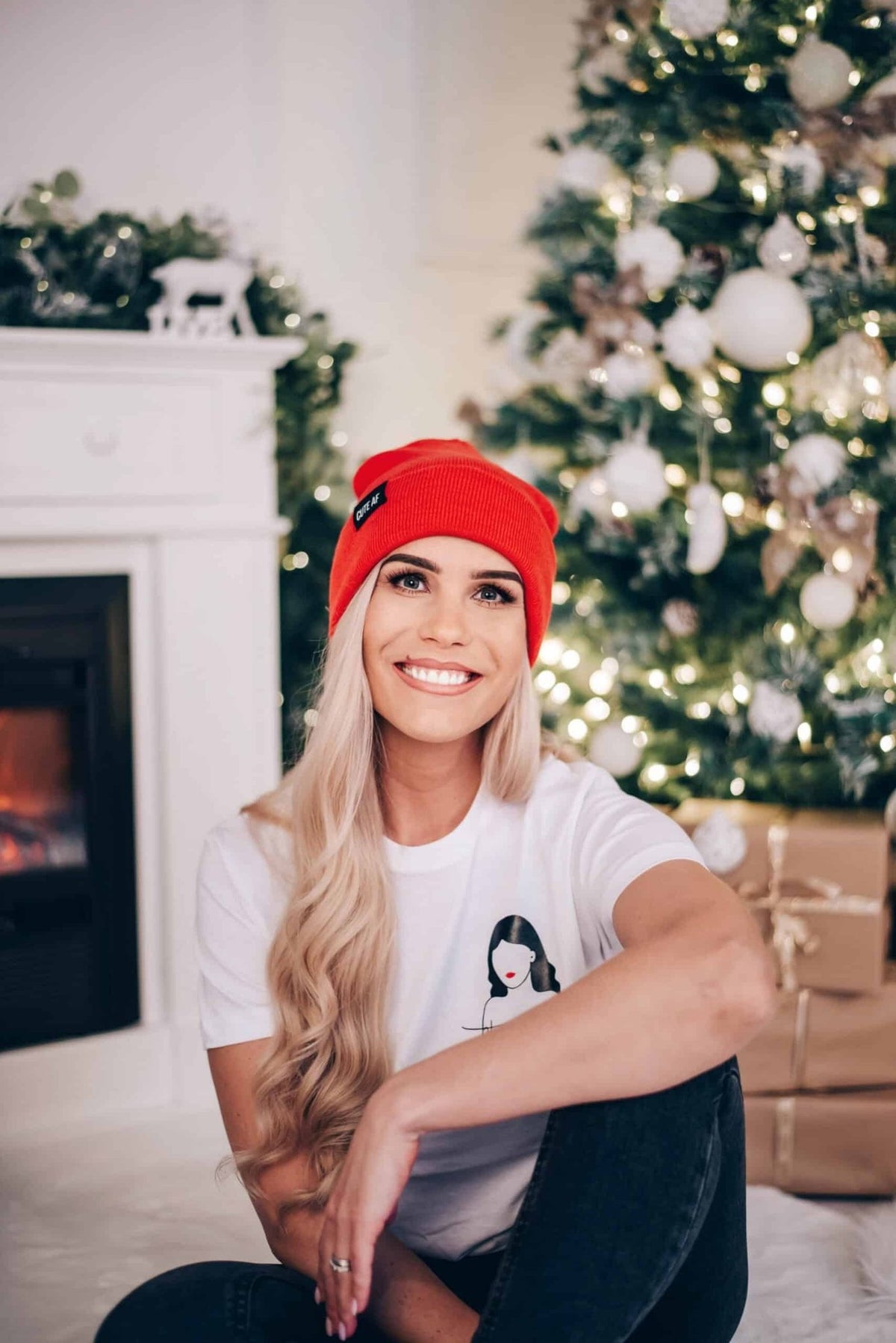 1. Woman sitting on floor in festive room wearing white 'Ballsy Christmas' T-shirt and red beanie, smiling in front of Christmas tree