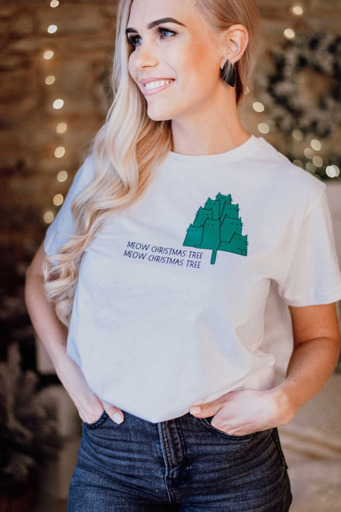 1. Woman wearing white T-shirt with green Christmas tree graphic and 'Meow Christmas Tree' text, smiling indoors with festive lights in background