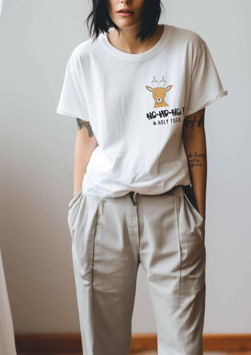 2. Woman in white Black Giraffe Brand T-shirt with reindeer graphic and 'Ho-ho-ho! & Holy Fuck' text, paired with beige pants in relaxed pose