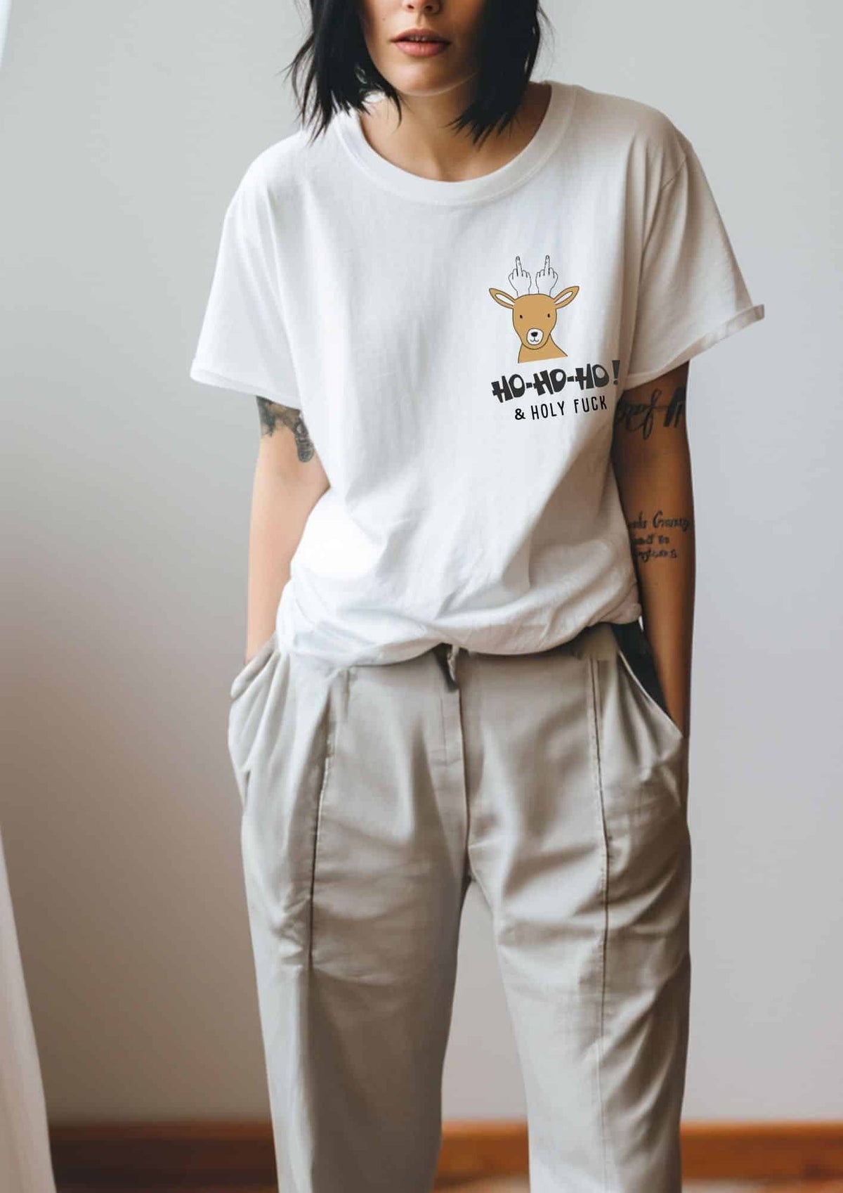 2. Woman in white Black Giraffe Brand T-shirt with reindeer graphic and 'Ho-ho-ho! & Holy Fuck' text, paired with beige pants in relaxed pose