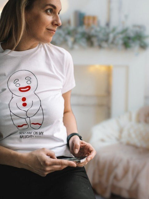 3. Woman wearing white T-shirt with gingerbread man print and 'You are on my naughty list' text, sitting in a cozy room